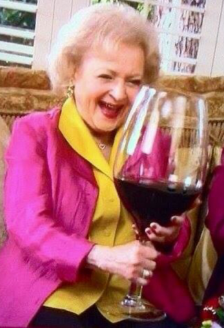 A woman joyfully holds an oversized wine glass filled with red wine, wearing a bright pink jacket over a yellow shirt.