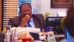 A man sitting at a desk, laughing and eating a snack, with a teddy bear in the background.